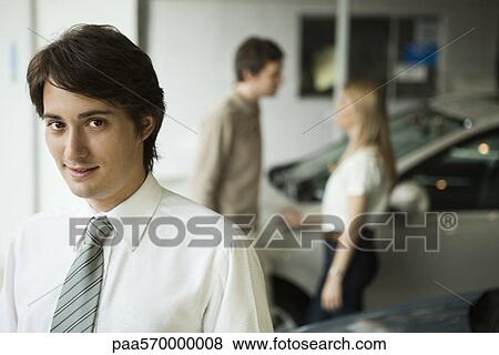Stock Photo - Car salesman in dealership showroom, potential buyers in background. Fotosearch
