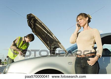 Stock Image - Woman making phone call while roadside assistance mechanic works to repair car. Fotosearch