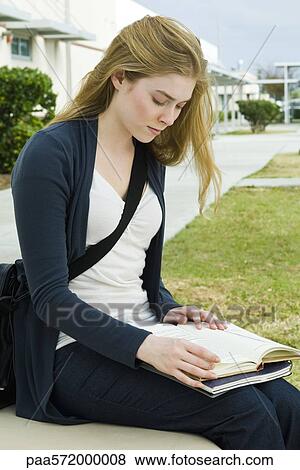 Stock Photo - Female college student sitting on bench reading book on campus. Fotosearch