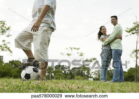 Boy playing with soccer ball, parents watching and embracing in background View Large Photo Image Stock Photo - Boy playing with soccer ball, parents watching and embracing in background. Fotosearch