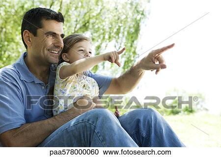 Stock Image - Father holding young daughter on lap outdoors, both pointing and looking away. Fotosearch
