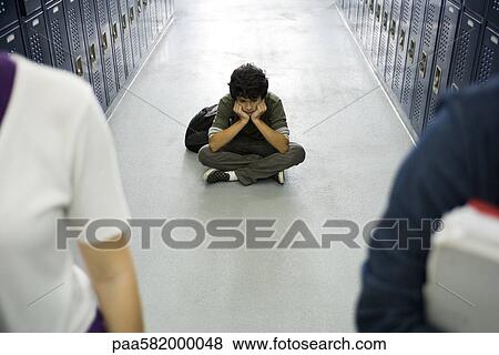 Stock Photo - Teenage boy sitting sullenly on floor of school corridor, classmates in foreground. Fotosearch