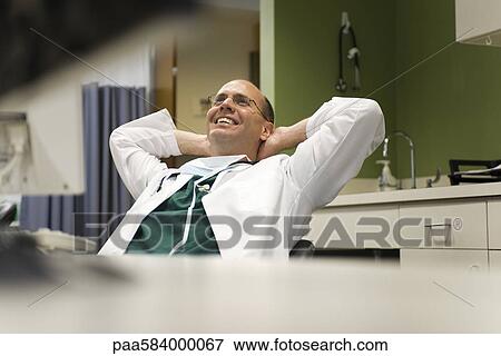 Stock Photo - Doctor leaning back in chair with hands behind head, smiling. Fotosearch
