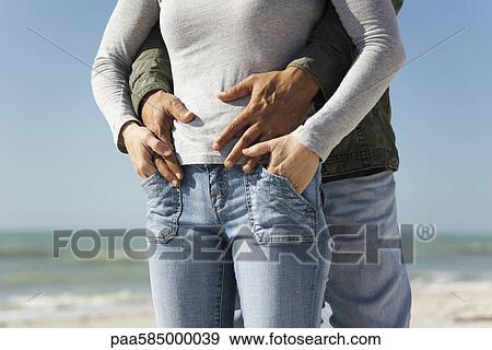 Stock Photo - Couple standing together at the beach, man embracing woman from behind. Fotosearch
