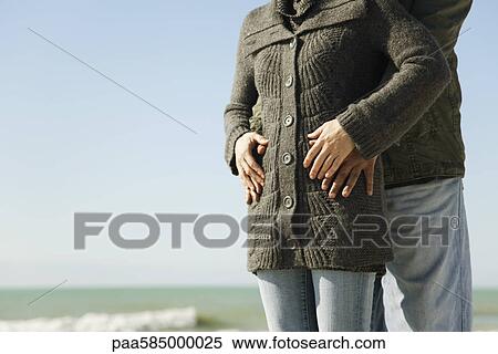 Stock Photography - Couple standing together at the beach, man embracing woman from behind. Fotosearch