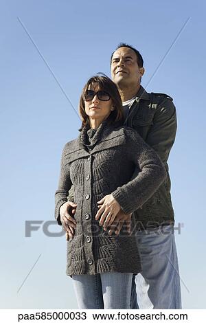 Couple standing together outdoors, man embracing woman from behind View Large Photo Image Stock Image - Couple standing together outdoors, man embracing woman from behind. Fotosearch