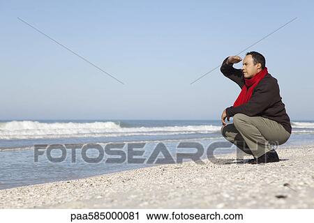 Stock Image - Man crouching on beach, looking at ocean. Fotosearch