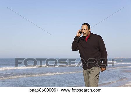 Stock Photo - Man talking on cell phone while walking on beach. Fotosearch