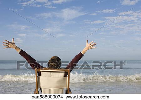 Picture - Male sitting in chair on beach with arms raised, rear view. Fotosearch
