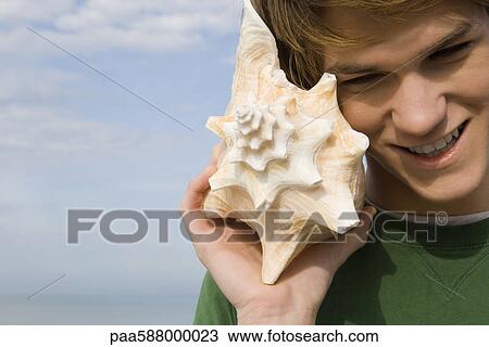 Stock Image - Young male listening to conch shell. Fotosearch