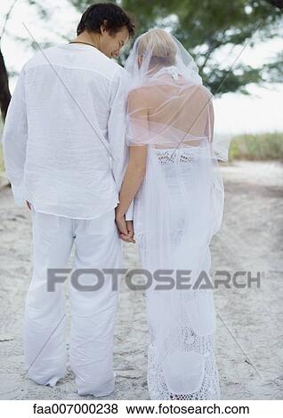 Stock Photo - Bride and groom walking on beach, holding hands, rear view. Fotosearch