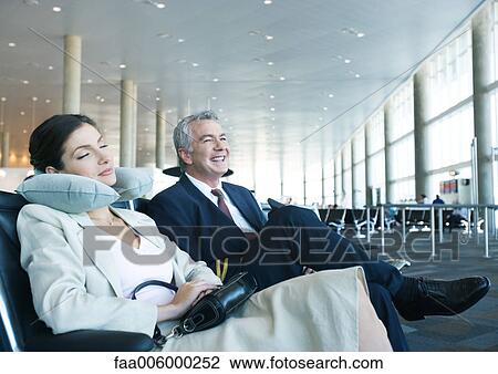 Stock Image - Business travelers sitting in airport lounge, woman napping with neck pillow next to smiling man. Fotosearch