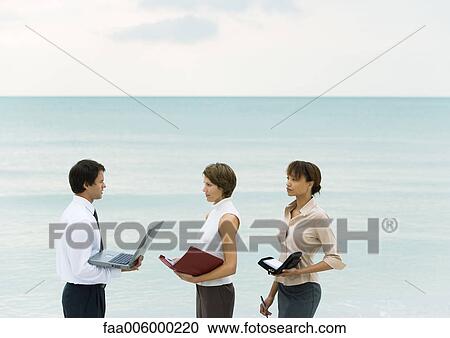 Businessman holding laptop, facing women holding agendas, sea in background View Large Photo Image Stock Image - Businessman holding laptop, facing women holding agendas, sea in background. Fotosearch