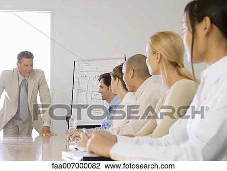 Businessman leaning on edge of conference table, speaking to team View Large Photo Image Stock Image - Businessman leaning on edge of conference table, speaking to team. Fotosearch