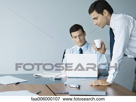 Stock Photo - Businessman leaning on table with cup in hand while colleague uses laptop. Fotosearch