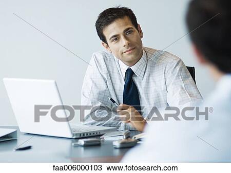 Businessman sitting at desk looking at second man View Large Photo Image Stock Image - Businessman sitting at desk looking at second man. Fotosearch