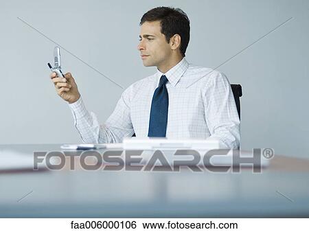 Businessman sitting at desk looking at cell phone View Large Photo Image Stock Photograph - Businessman sitting at desk looking at cell phone. Fotosearch