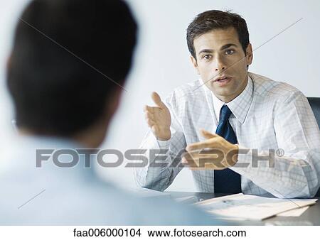 Businessman sitting at desk speaking to second man View Large Photo Image Picture - Businessman sitting at desk speaking to second man. Fotosearch