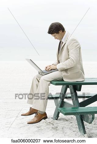 Businessman sitting on edge of picnic table at beach, using laptop View Large Photo Image Stock Photo - Businessman sitting on edge of picnic table at beach, using laptop. Fotosearch