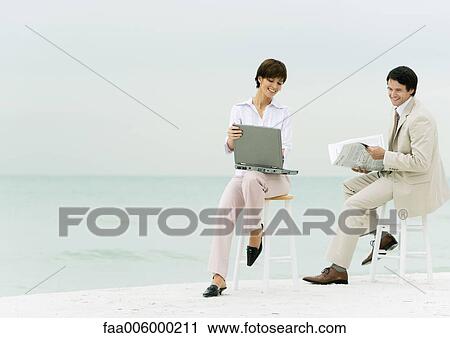 Stock Image - Businesspeople sitting on stools on beach, one using laptop, another reading newspaper. Fotosearch