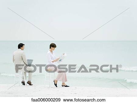 Stock Photo - Businesspeople sitting on stools on beach, one using laptop, another reading newspaper. Fotosearch