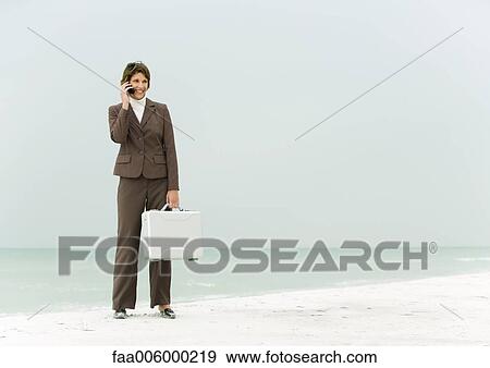 Businesswoman standing on beach holding briefcase, using cell phone View Large Photo Image Stock Photo - Businesswoman standing on beach holding briefcase, using cell phone. Fotosearch