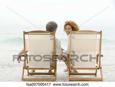 Stock Photo - Couple sitting in beach chairs, woman turning around, looking at camera. Fotosearch