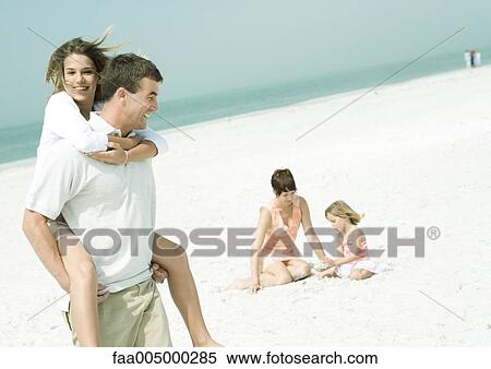 Stock Photography - Family at the beach, man carrying teenage daughter piggyback while mother sits on sand with daughter. Fotosearch
