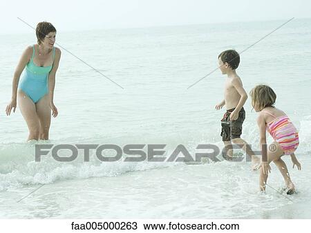Stock Image - Family at the beach, mother standing in the waves looking at children coming into water. Fotosearch