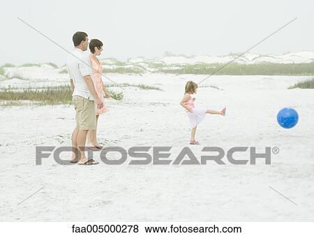 Stock Photo - Family at the beach, parents watching as little girl kicks ball. Fotosearch