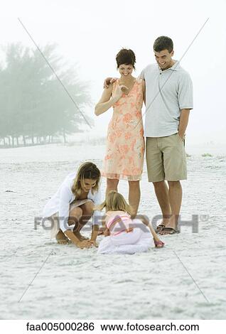 Stock Photograph - Family at the beach, parents watching daughters play in sand. Fotosearch