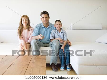 Stock Photograph - Father and two children sitting on sofa, pointing remote control. Fotosearch