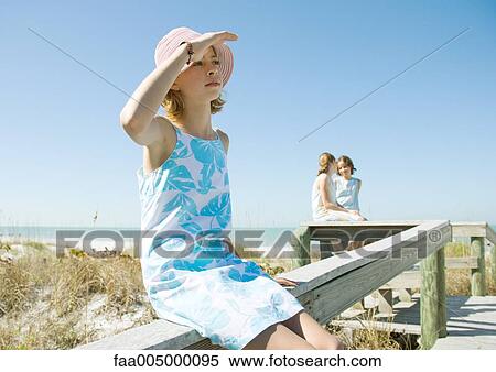 Stock Photography - Girl sitting on railing of beach walkway, shading eyes. Fotosearch