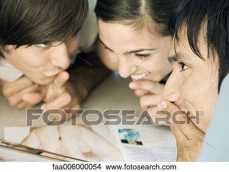 Picture - Group of young adult friends leaning over magazine together, sharing secret, close-up. Fotosearch