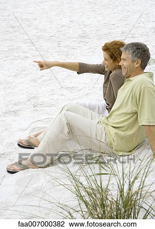 Stock Image - Mature couple sitting on beach, side view. Fotosearch