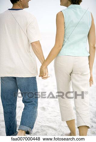 Stock Photo - Mature couple walking hand in hand on beach, rear view. Fotosearch