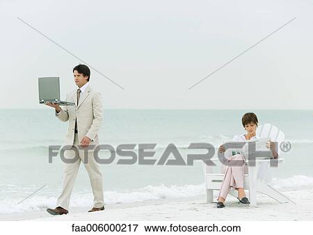 On beach, businessman carrying laptop, walking by woman reading newspaper in chair View Large Photo Image Stock Photo - On beach, businessman carrying laptop, walking by woman reading newspaper in chair. Fotosearch