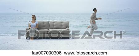 Stock Photograph - On beach, woman sitting on sofa with eyes closed while man runs by behind her. Fotosearch