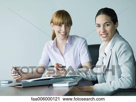 Stock Image - Two businesswomen sitting at desk, smiling at camera. Fotosearch
