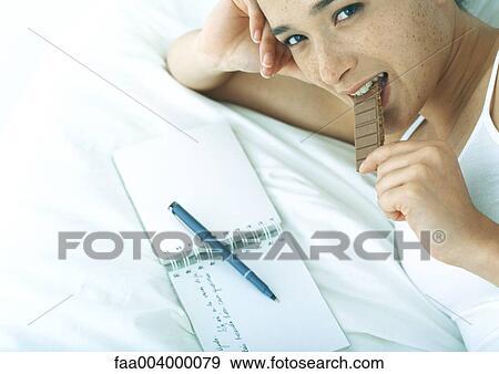 Woman lying on bed in front of open notebook, eating chocolate View Large Photo Image Stock Photo - Woman lying on bed in front of open notebook, eating chocolate. Fotosearch