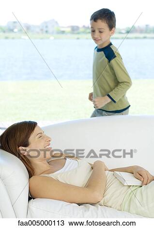 Woman lying on sofa while son runs around her View Large Photo Image Stock Image - Woman lying on sofa while son runs around her. Fotosearch