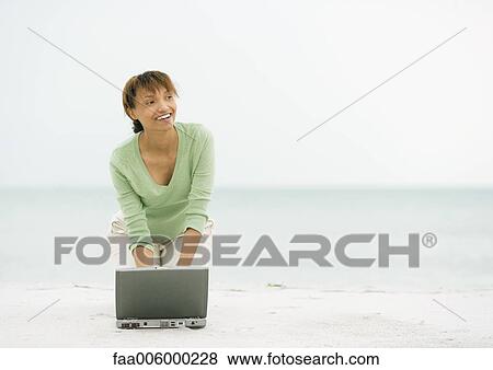 Stock Photo - Woman on beach, kneeling on sand with laptop. Fotosearch
