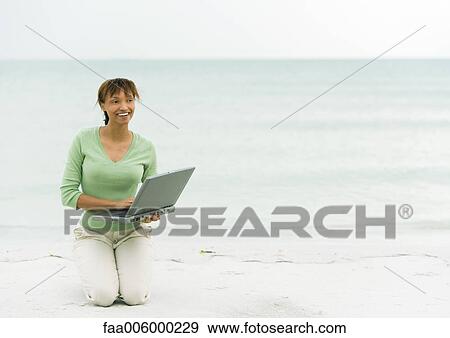 Stock Photo - Woman on beach, kneeling on sand with laptop. Fotosearch