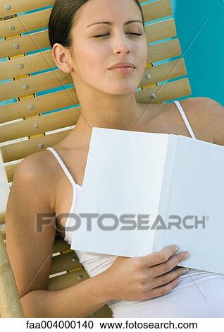 Stock Image - Woman sleeping in lounge chair, holding book on chest. Fotosearch