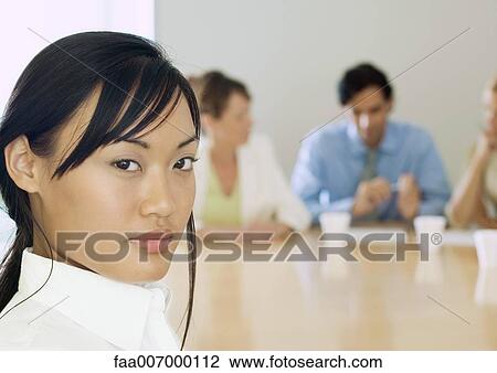 Stock Image - Young businesswoman, colleagues in meeting in background. Fotosearch