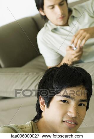 Stock Photo - Young man smiling, friend lying on couch in background. Fotosearch