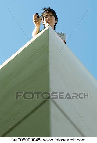 Young man using cell phone on top of building, low angle view View Large Photo Image Stock Photography - Young man using cell phone on top of building, low angle view. Fotosearch