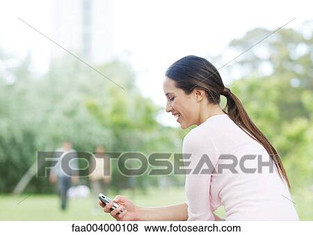 Stock Photo - Young woman checking cell phone in city park. Fotosearch