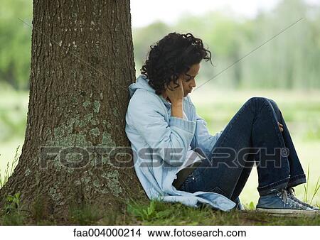 Picture - Young woman sitting on ground, leaning against tree. Fotosearch