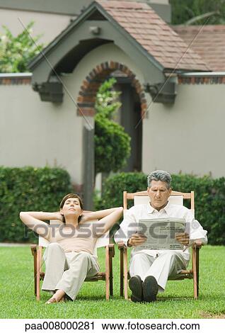 Stock Image - Couple sitting in deckchairs on lawn, man reading newspaper and woman napping. Fotosearch
